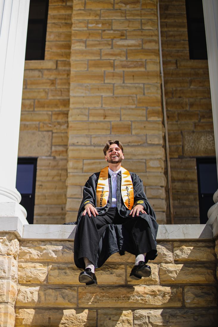 A graduate in cap and gown sitting on a stone ledge, laughing, in front of a sandstone wall