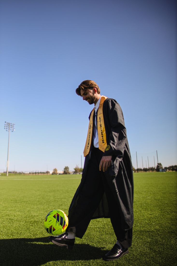 A graduate in cap and gown looking down at a soccer ball on the pitch