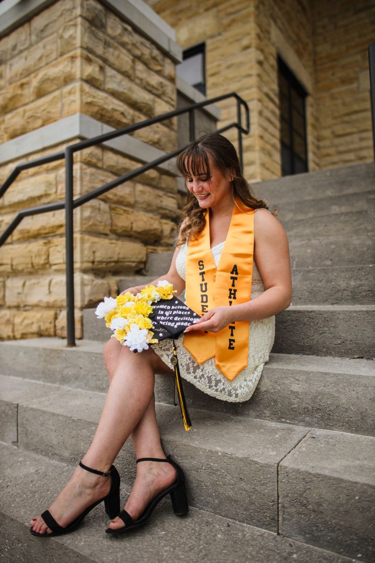 A graduate in a Student Athlete stole sitting on stone steps holding a decorated cap